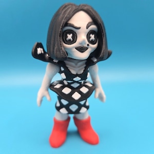 May include: A small figurine with black hair, pale skin, and button eyes shaped like white crosses. The figure wears a black and white diamond-patterned dress with a flared peplum waist and bright red boots. The character has a small black dot on the cheek.