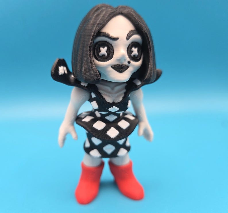 May include: A small figurine with black hair, pale skin, and button eyes shaped like white crosses. The figure wears a black and white diamond-patterned dress with a flared peplum waist and bright red boots. The character has a small black dot on the cheek.