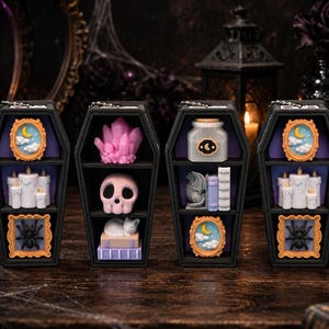 May include: Four black coffin-shaped shelves featuring miniature decorative items. The shelves contain small framed moon and cloud art, white pillar candles, black spiders, a pink crystal cluster, a pink skull, a white cat, and a potion bottle.