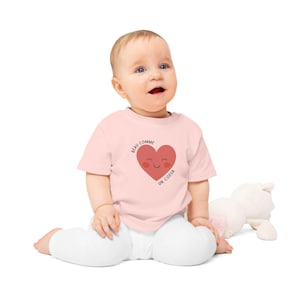 Peut inclure: Un bébé portant un t-shirt rose clair avec un cœur rouge souriant. Le texte "BEAU COMME UN COEUR" est imprimé autour du cœur. Le bébé est assis sur une surface blanche.