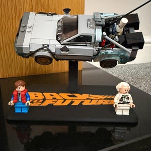 May include: A Lego DeLorean time machine model, suspended above a black base with the words "BACK TO THE FUTURE" in orange. Two Lego figures, one in a red vest and the other in a white coat, stand on the base. The car is silver and grey.