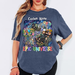 May include: A navy blue t-shirt featuring a colorful graphic with the text "Custom Name" above a design with various cartoon characters and the text "2026". The words "EPIC UNIVERSE" are displayed in colorful letters below the graphic.