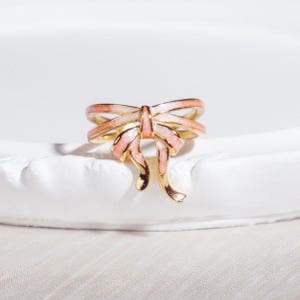Può includere: Un anello dorato con un design a fiocco con dettagli in smalto rosa e bianco. I nastri del fiocco si incurvano verso il basso, creando un aspetto delicato e femminile. L'anello è presentato su uno sfondo bianco, evidenziando i suoi dettagli.