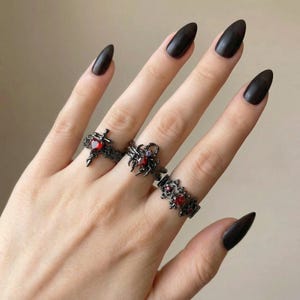 May include: Three black rings with red gemstone accents are displayed on a hand with long, dark nails. The rings feature gothic and ornate designs, including a cross, a spider, and a floral pattern, creating a dark and edgy aesthetic.
