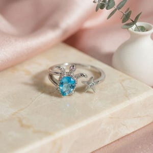 May include: A silver-toned open ring featuring a sea turtle motif with a pear-shaped blue gemstone set in the shell and a small starfish accent on the opposite end. The band is crafted from polished metal with delicate detailing on the sea creature.