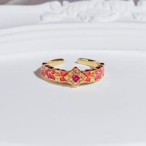 Puede incluir: Anillo ajustable dorado con una banda de esmalte rosa. El anillo presenta un diseño central en forma de diamante con una gema roja y cristales transparentes circundantes. La banda tiene un patrón repetitivo de esmalte rosa y naranja.