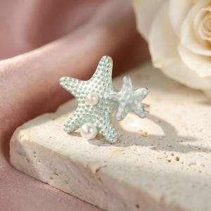 May include: A ring featuring two connected starfish shapes in a pale mint green color with a textured, dotted surface. The larger starfish is accented with two small, round white pearls embedded into its surface, creating a detailed design.