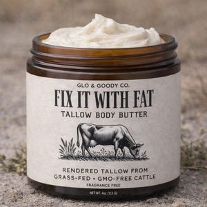 Puede incluir: Un tarro de manteca corporal de sebo con un recipiente de vidrio marrón y una etiqueta blanca. La etiqueta dice "FIX IT WITH FAT" y presenta una ilustración de vaca. El tarro contiene una manteca corporal cremosa, blanquecina. Peso neto 113 g.