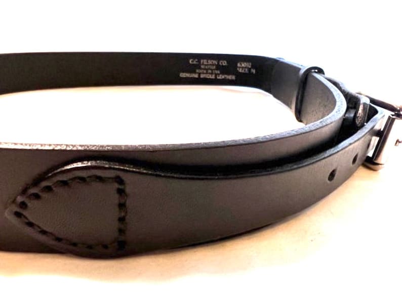 K&ouml;nnte beinhalten: Nahaufnahme eines schwarzen Lederg&uuml;rtels mit silberner Schnalle. Der G&uuml;rtel hat eine gen&auml;hte Lederschlaufe und ist mit "Genuine Bridle Leather" und "Made in USA" gekennzeichnet. Der G&uuml;rtel ist etwa 3,18 cm breit.