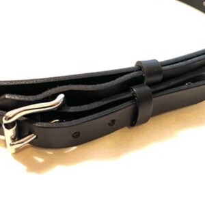 K&ouml;nnte beinhalten: Nahaufnahme eines schwarzen Lederg&uuml;rtels mit silberfarbener Schnalle. Der G&uuml;rtel hat ein klassisches Design mit einer Dornschlie&szlig;e und mehreren L&ouml;chern zur Weitenregulierung. Das Leder wirkt glatt und ist f&uuml;r den t&auml;glichen Gebrauch geeignet.