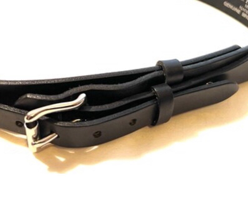 K&ouml;nnte beinhalten: Nahaufnahme eines schwarzen Lederg&uuml;rtels mit silberfarbener Schnalle. Der G&uuml;rtel hat ein klassisches Design mit einer Dornschlie&szlig;e und mehreren L&ouml;chern zur Weitenregulierung. Das Leder wirkt glatt und ist f&uuml;r den t&auml;glichen Gebrauch geeignet.