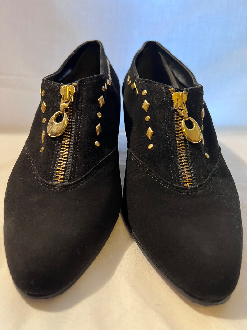 May include: A pair of black suede ankle boots with gold-tone zippers and studs. The boots have a pointed toe and a low heel.