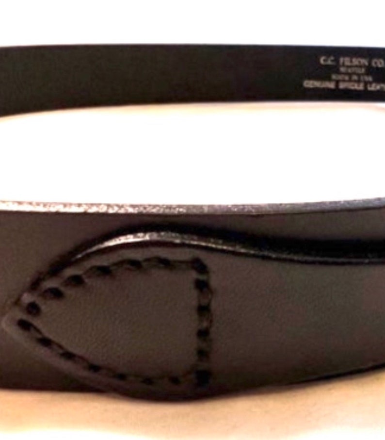 K&ouml;nnte beinhalten: Nahaufnahme eines schwarzen Lederg&uuml;rtels mit einem gen&auml;hten Design in der N&auml;he der Schnalle. Der G&uuml;rtel tr&auml;gt den Text "C.C. FILSON CO. SEATTLE MADE IN USA GENUINE BRIDLE LEATHER".