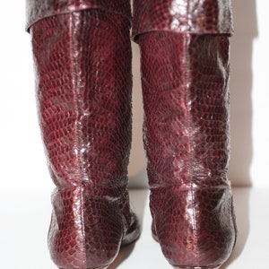 May include: A pair of burgundy knee-high boots with a pointed toe and a low heel. The boots are made of a textured leather that resembles snakeskin.