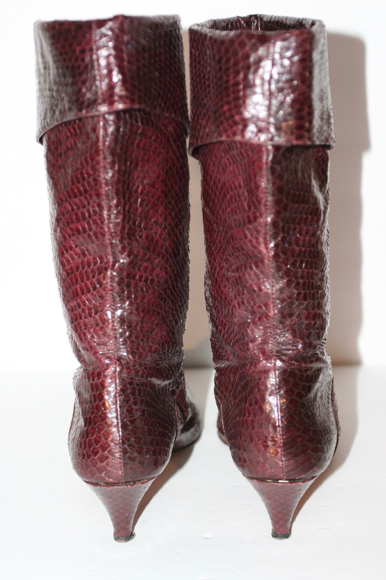May include: A pair of burgundy knee-high boots with a pointed toe and a low heel. The boots are made of a textured leather that resembles snakeskin.