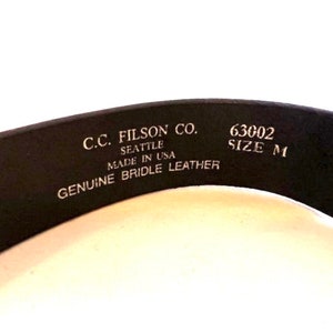 K&ouml;nnte beinhalten: Nahaufnahme eines schwarzen Lederg&uuml;rtels mit dem Text "C.C. Filson Co. Seattle Made in USA Genuine Bridle Leather." Der G&uuml;rtel hat auch die Gr&ouml;&szlig;e "M" und die Nummer "63002".