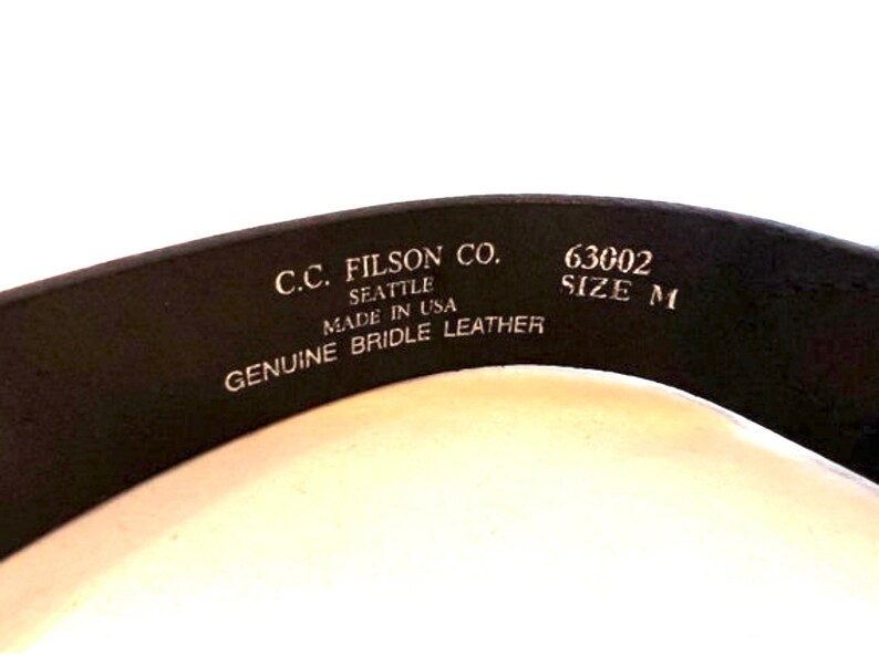 K&ouml;nnte beinhalten: Nahaufnahme eines schwarzen Lederg&uuml;rtels mit dem Text "C.C. Filson Co. Seattle Made in USA Genuine Bridle Leather." Der G&uuml;rtel hat auch die Gr&ouml;&szlig;e "M" und die Nummer "63002".
