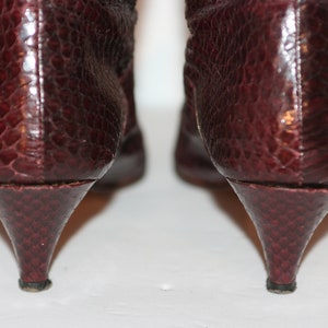 May include: A pair of burgundy snakeskin heeled boots with pointed toes. The boots are shown from the back, highlighting the heels.