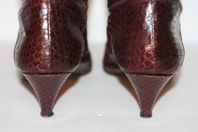 May include: A pair of burgundy snakeskin heeled boots with pointed toes. The boots are shown from the back, highlighting the heels.