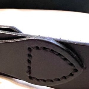 K&ouml;nnte beinhalten: Nahaufnahme einer schwarzen Lederschlaufe mit einer gen&auml;hten Umrandung. Die Schlaufe hat eine abgerundete rechteckige Form und ist am G&uuml;rtel befestigt. Das Leder scheint glatt zu sein und die N&auml;hte sind sichtbar.