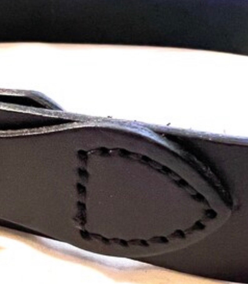 K&ouml;nnte beinhalten: Nahaufnahme einer schwarzen Lederschlaufe mit einer gen&auml;hten Umrandung. Die Schlaufe hat eine abgerundete rechteckige Form und ist am G&uuml;rtel befestigt. Das Leder scheint glatt zu sein und die N&auml;hte sind sichtbar.