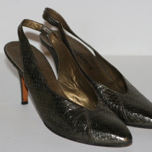 Puede incluir: Un par de tacones de slingback metálicos plateados con punta puntiaguda. Los tacones están hechos de un material texturizado y tienen una fina correa que rodea el tobillo.