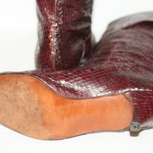 May include: A pair of burgundy leather boots with a snakeskin pattern. The boots have a low heel and a pointed toe. The boots are shown from the side, with the sole of one boot visible.