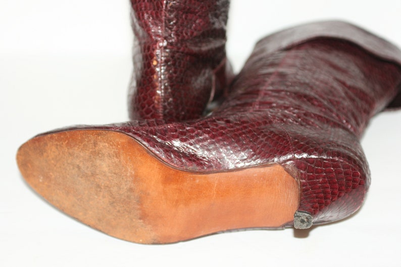 May include: A pair of burgundy leather boots with a snakeskin pattern. The boots have a low heel and a pointed toe. The boots are shown from the side, with the sole of one boot visible.