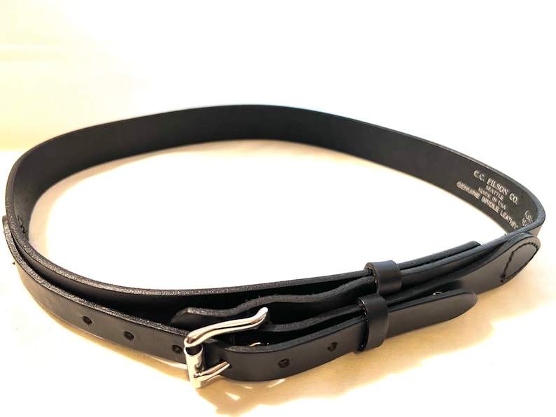 K&ouml;nnte beinhalten: Ein schwarzer Lederg&uuml;rtel mit silberner Schnalle. Der G&uuml;rtel ist aus echtem Zaumleder gefertigt und tr&auml;gt den Aufdruck "C.C. Filson Co. Seattle Made in USA". Der G&uuml;rtel ist etwa 2,5 cm breit.