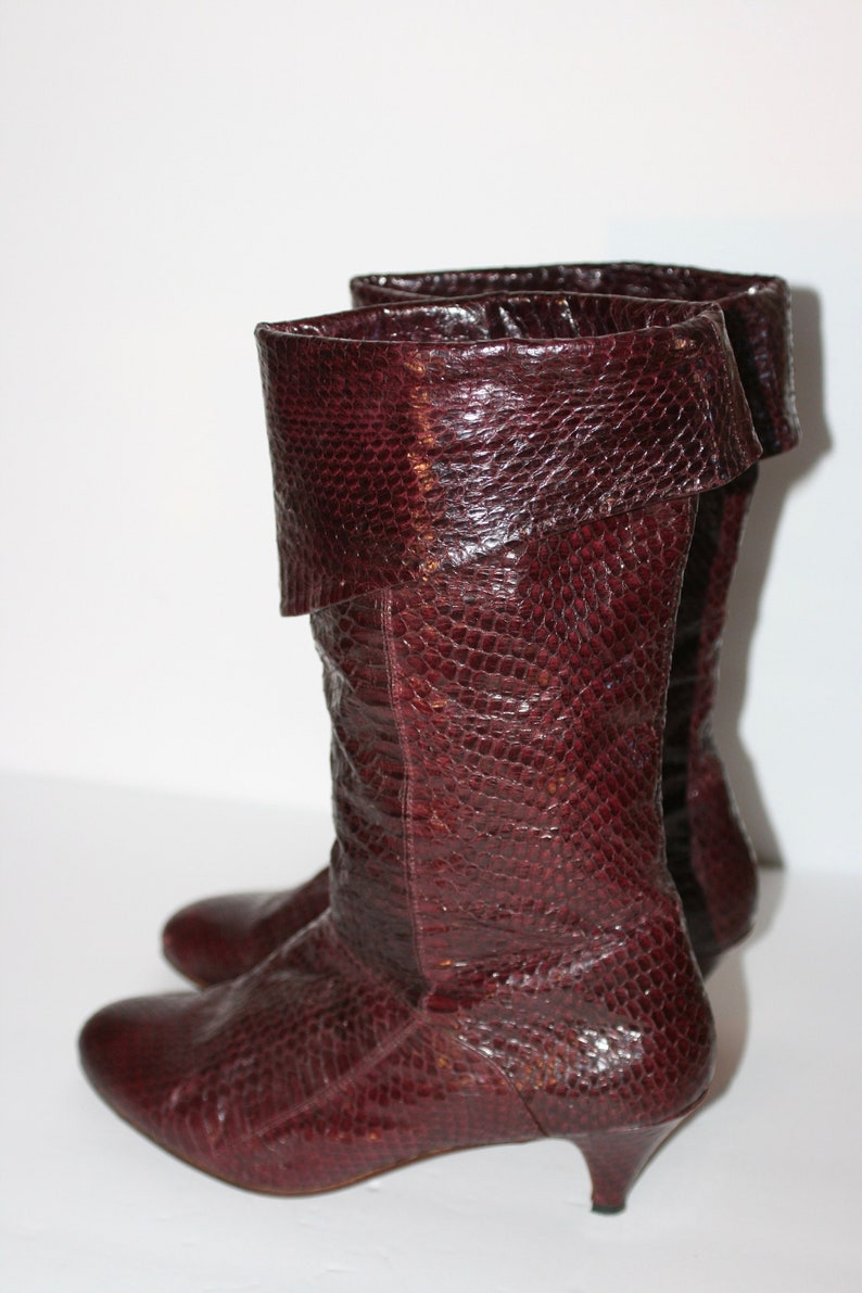 May include: A pair of burgundy leather boots with a snakeskin pattern. The boots have a low heel and a fold-over cuff.