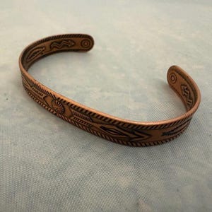 May include: A copper cuff bracelet featuring an open design and a series of engraved geometric patterns, including arrows, sunbursts, and zigzag lines. The metal has a warm, reddish-brown tone with dark oxidized accents highlighting the engravings.