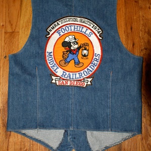 May include: A blue denim vest with a colourful patch featuring a cartoon character holding a lantern. The patch reads "NMRA Silver Gate 1974 Foothills Model Railroaders San Diego."