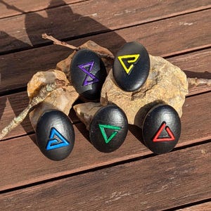 Può includere: Sei pietre nere di forma ovale con simboli geometrici colorati. I simboli sono delineati in blu, verde, rosso, giallo e viola. Le pietre sono su una roccia e una superficie di legno.