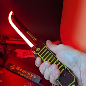 Może przedstawiać: Czarny i żółty nóż Starsys Kelvin K-4 z czerwoną świecącą klingą. Rękojeść ma wzór w paski. Nóż trzymany jest w dłoni, z tekstem "Starsys Kelvin K-4" widocznym na oddzielnym elemencie.