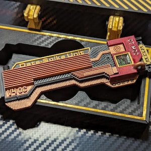 May include: A rectangular Data-Spike Unit prop with a copper-colored ribbed body, a red section featuring the text OC MOD and STARBYS, and a black base. The unit is housed in a black and gold storage case with a carbon fiber pattern on the interior.
