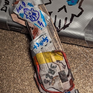 May include: A handmade, mixed-media art piece featuring a skull drawing with blue accents and the text "Ø-DA". The piece incorporates various materials, including tape and what appears to be electronic components, with handwritten text and doodles.
