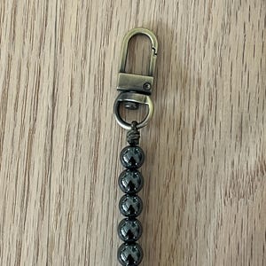 May include: A keychain featuring a series of dark gray, glossy beads strung together. The beads are connected to a bronze-colored clasp. The background is a light brown wooden surface.