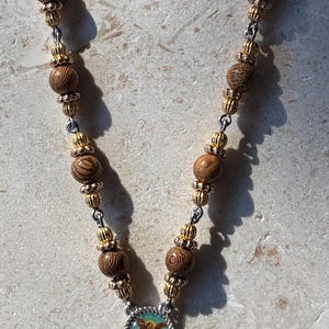 May include: A rosary necklace featuring brown wood beads, gold-tone metal spacers, and silver-tone accents. It includes a circular medal depicting a religious figure, a single green stone bead, and a silver-tone crucifix with a red enamel inlay.