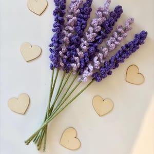 May include: A handmade bouquet of crocheted lavender flowers in shades of purple and lavender, with green stems. Several small, wooden heart shapes are scattered around the flowers. The arrangement is on a white background.