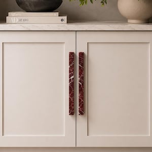 May include: A pair of vertical, rectangular cabinet handles made of deep red marble with white veining, mounted on light beige double cabinet doors. The handles feature a smooth, polished finish and a straight, elongated design.