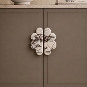 May include: Close-up of a cabinet with a pair of flower-shaped marble door pulls. The pulls are white with dark gray and brown veining. The cabinet doors are a muted brown color. A book titled "Picasso" is visible.