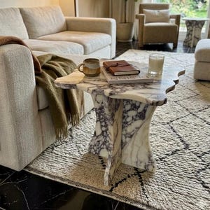 May include: A living room scene featuring a white sofa with a brown throw blanket and a green blanket. A marble coffee table with a unique shape holds a mug, a book, and a candle. A patterned rug and a floor lamp complete the cozy setting.