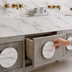 May include: A bathroom vanity featuring a white marble countertop with gray veining, brass wall-mounted faucets, and gray wood cabinets. The cabinet doors and drawers are accented with circular white marble pulls split into two halves.