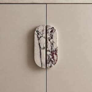 May include: Close-up of a cabinet with two marble door pulls. The pulls are a light gray with black and burgundy veining. The cabinet doors are a neutral beige color. The pulls are oval shaped.