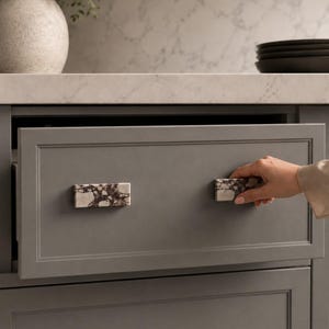 May include: Gray kitchen cabinet drawer with two rectangular marble cabinet pulls. The drawer is being opened by a person. The marble pulls have a white base with dark brown veining.
