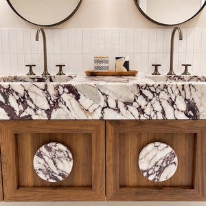 May include: A double vanity featuring a white and purple marble countertop with two integrated sinks, brass faucets, and two round wooden cabinet doors accented with circular marble inlays, positioned below two round mirrors and white wall tiles.
