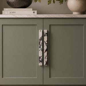 May include: Sage green cabinet doors with marble-patterned handles. The cabinet doors are set against a neutral background. A decorative bowl and books are on the countertop. A vase with greenery is also visible.