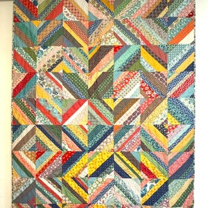 May include: A colorful patchwork quilt with a geometric design. The quilt features a variety of fabrics in shades of red, blue, yellow, green, and brown. The design is made up of diagonal stripes and squares, creating a visually interesting pattern. The quilt is hanging on a wall.