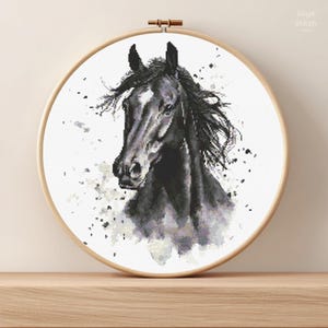 Op de afbeelding: Een zwart-wit kruissteekborduurwerk van een paardenhoofd, omlijst in een houten hoepel. Het paard heeft een donkere manen en een witte bles op zijn gezicht. De achtergrond heeft spatten.
