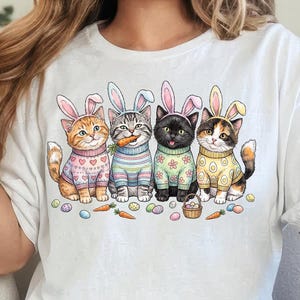 May include: White t-shirt featuring a colorful graphic of four cats dressed in Easter bunny outfits. The cats are wearing sweaters and bunny ears, surrounded by Easter eggs and carrots. The design is whimsical and festive.